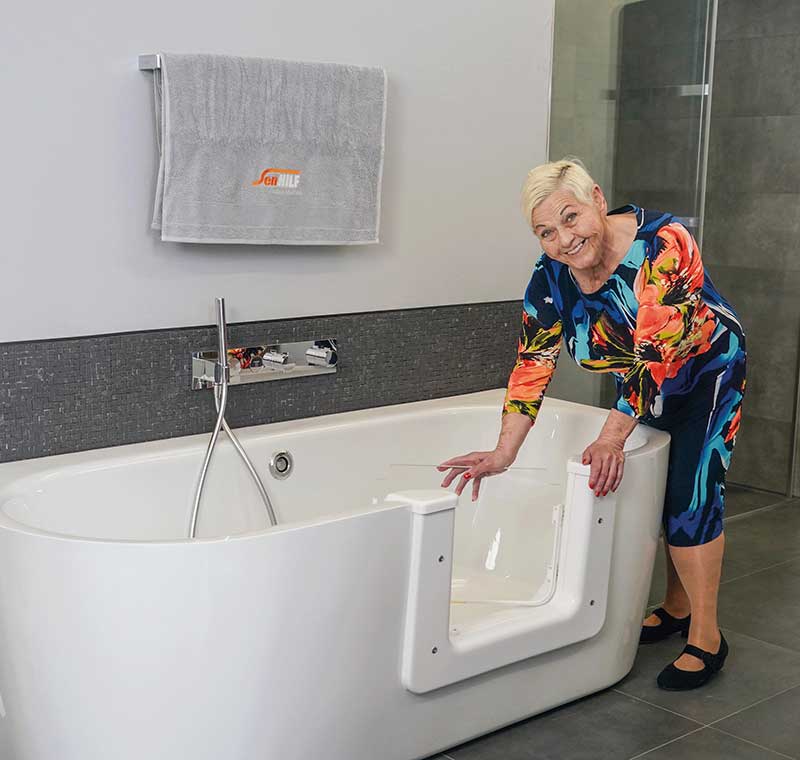 Eine Frau der Generation 50plus in einem farbenfrohen Kleid lächelt, während sie neben einer weißen, begehbaren Badewanne in einem modernen Badezimmer steht und ihre Hand auf die Wannentür stützt. Ein Handtuch hängt fein säuberlich an der Wand hinter ihr. Meckel-Bad Plauen