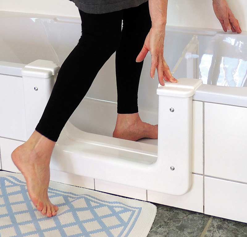 Eine Person der Generation 50plus steigt mit einem Bein innerhalb und einem Bein außerhalb der Wanne über eine weiße Sicherheitstreppe in eine Badewanne. Auf dem gefliesten Boden liegt eine blau-weiße Matte. Meckel-Bad Plauen