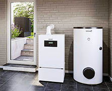 Ein modernes Wärmepumpensystem von Heizung und Wärme ist in einem Innenraum in der Nähe einer Glastür installiert, mit einem zylindrischen Wassertank und einer rechteckigen Wärmepumpeneinheit auf einem gefliesten Boden vor einer Ziegelwand. Meckel-Bad Plauen