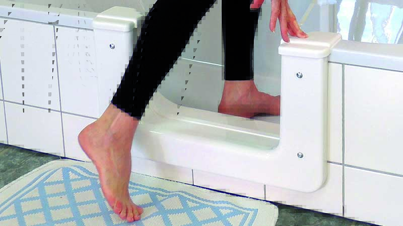 Eine Person steigt in eine Badewanne, indem sie einen Wannenausschnitt in ihrem Bad und ihrer Heizung benutzt, der einen niedrigeren Einstiegspunkt für einen leichteren Zugang schafft. Ein Fuß befindet sich in der Wanne, während der andere über den Ausschnitt auf eine blau-weiße Badematte tritt. Meckel-Bad Plauen