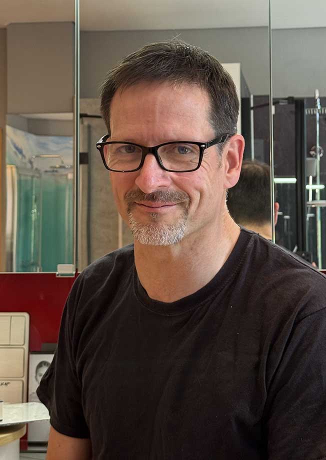 Herr Meckel mit kurzen braunen Haaren, Brille und einem graumelierten Spitzbart lächelt in einem schwarzen Hemd. Er steht in einem Raum, im Hintergrund sind Spiegel und verschiedene Gegenstände zu sehen, die mit „Bad und Heizung“ zu tun haben. Meckel-Bad Plauen