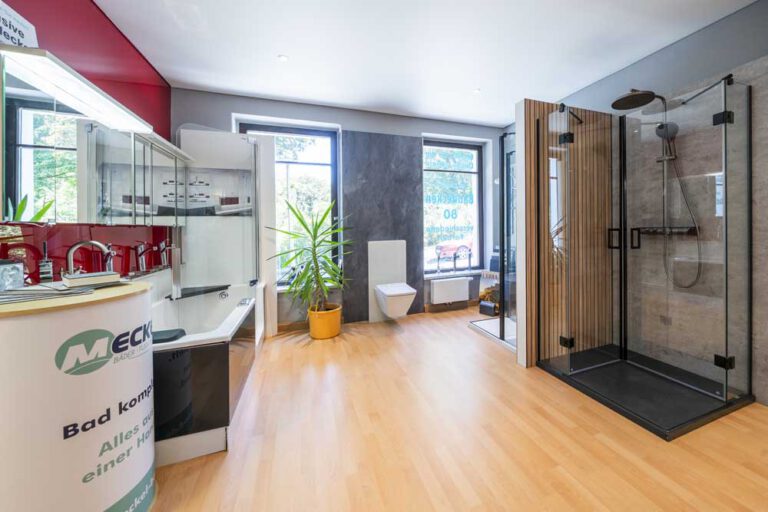 Moderner Badezimmer-Showroom mit Badewanne, Waschbecken, Toilette und Glasduschkabine – die perfekte Inspiration für Ihre nächste Badrenovierung. Große Fenster, ein Holzboden und eine Topfpflanze verleihen dem Raum Helligkeit. Auf der linken Seite ist eine Ausstellungstheke zu sehen. Meckel-Bad Plauen